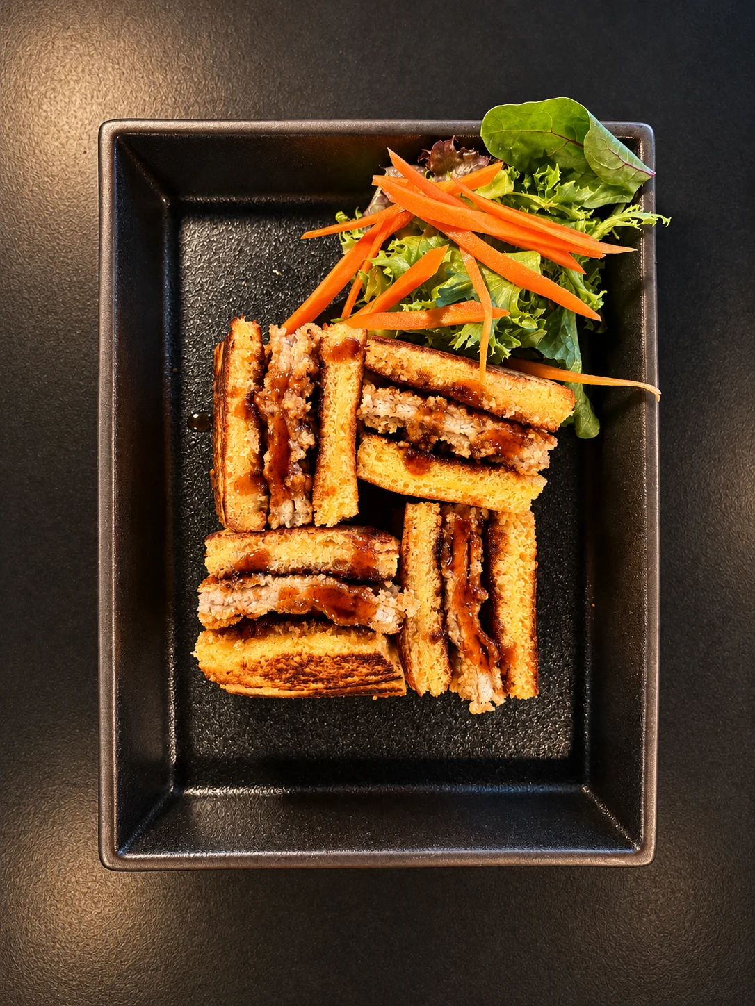 Four Katsu Sando sandwich pieces with sauce and mixed greens on a black platter