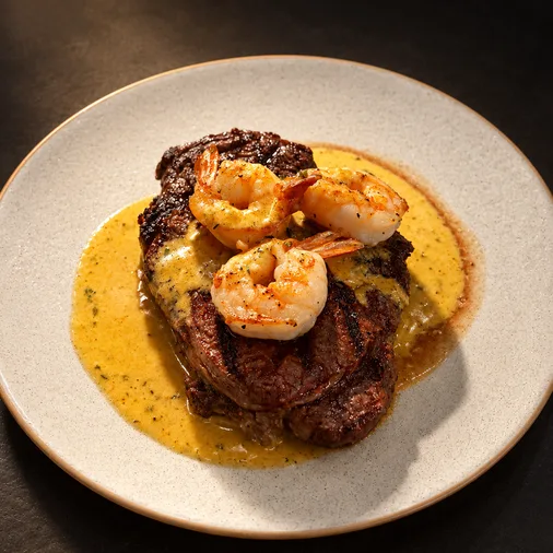 Ribeye topped with three grilled tiger shrimp and creamy sauce on a white plate