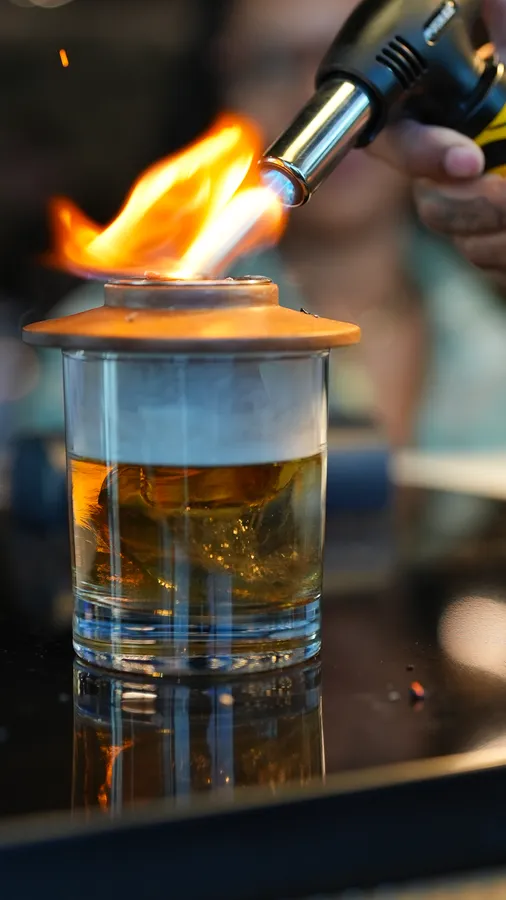 Smokey Old Fashion cocktail being infused with smoke beneath a copper lid and torch flame