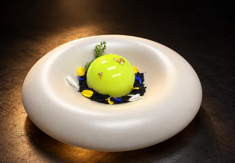 Green-glazed Yuzu Cheesecake with gold leaf, dark crumble, and edible flowers on a white plate